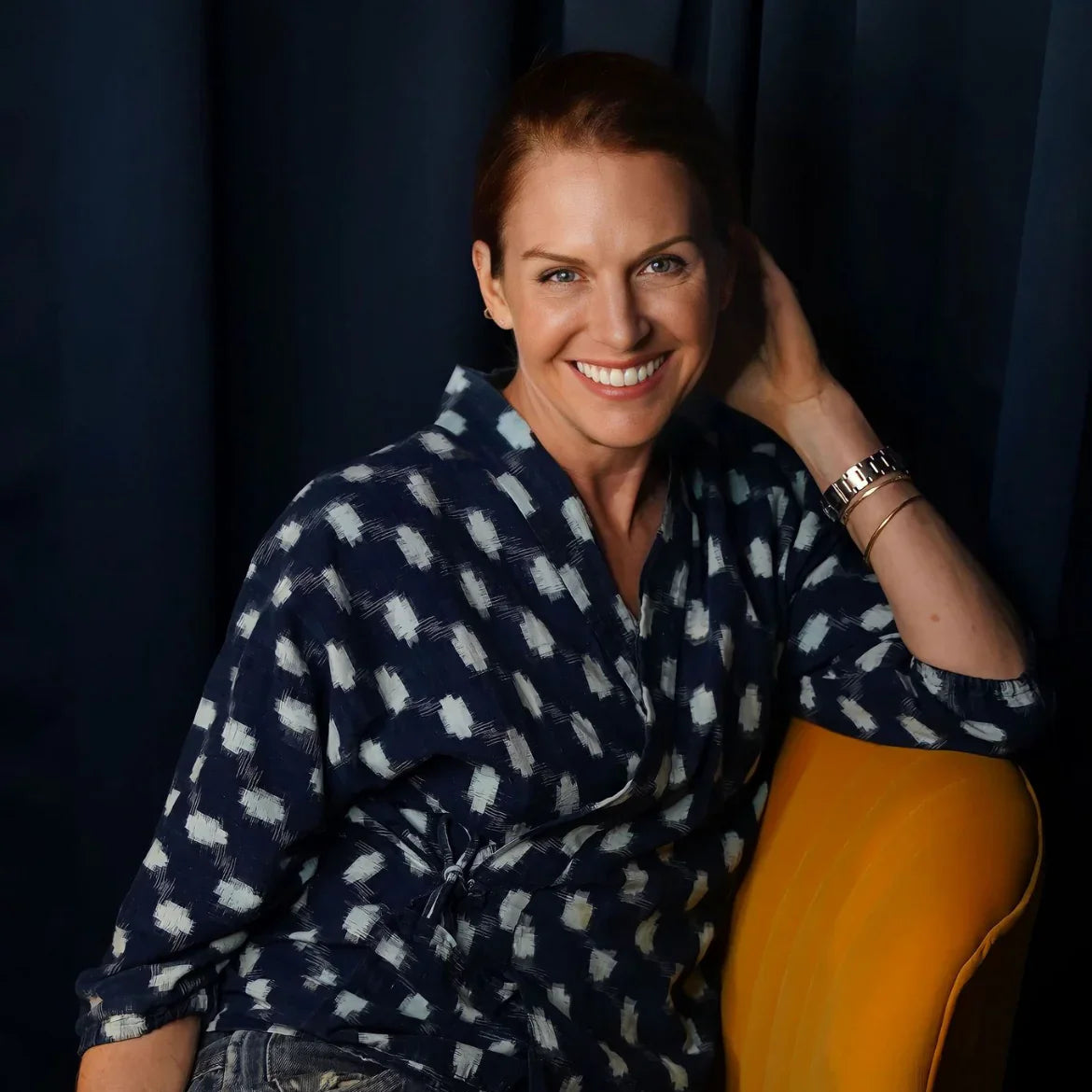 Smiling woman in patterned navy shirt and jeans sitting on yellow chair against dark backdrop