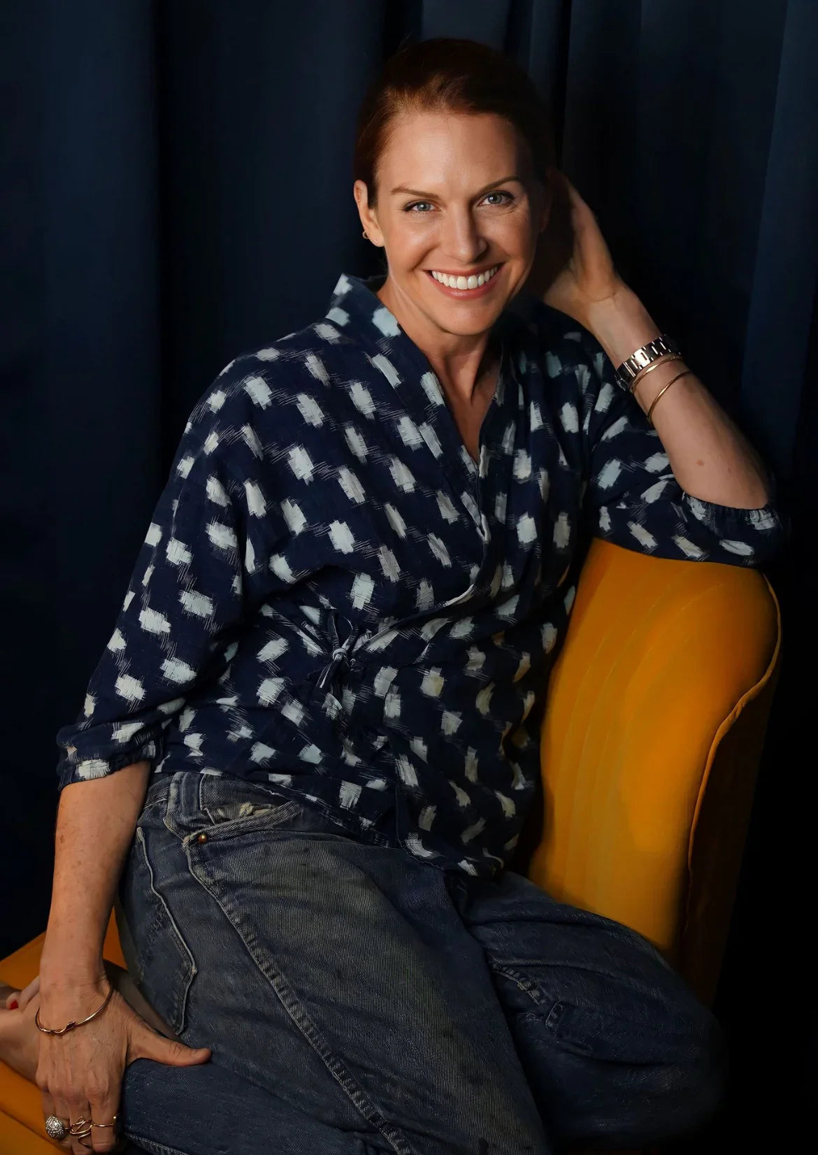 Smiling woman in patterned navy shirt and jeans sitting on yellow chair against dark backdrop